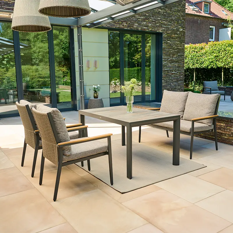 Modern eingerichteter Terrassenbereich mit einem rechteckigen Esstisch aus Holz und dunklem Metallgestell, flankiert von zwei gepolsterten Gartenstühlen und einer zweisitzigen Gartenbank. Unter dem Tisch liegt ein helles Outdoor Teppich. Im Hintergrund reflektieren große Glasfronten den Garten mit Bäumen und Hecken