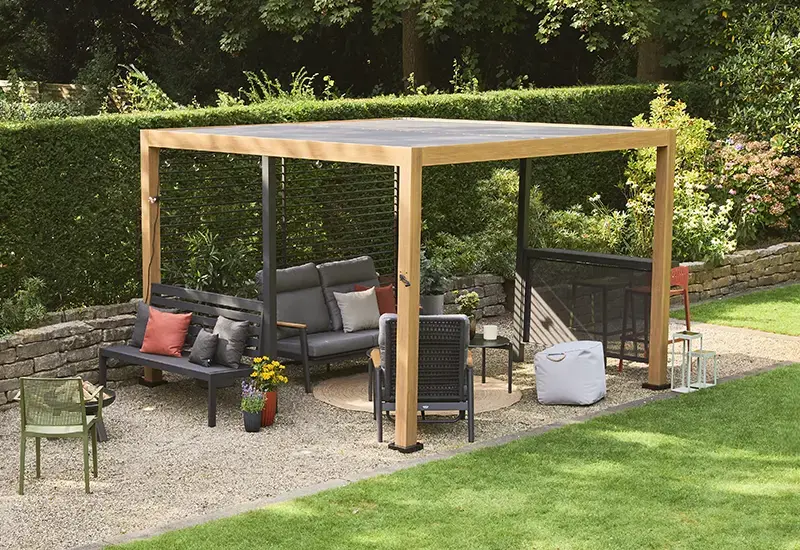 Moderne Pergola mit Gestell in Holzoptik und verstellbarem Lamellendach über einer gemütlichen Gartenlounge aus Sofas, Sesseln und Beistelltischen, umgeben von Grünpflanzen.