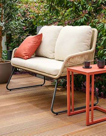 Zweier Outdoor Bank aus hellem Geflecht mit beigen Polstern und rotem Kissen neben einem roten Beistelltisch auf einer Holzterrasse im Garten.