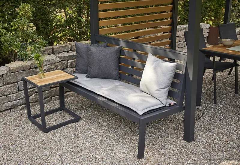 Outdoor Sitzbank aus Metall mit hellen Polstern und Kissen neben einem kleinen Beistelltisch vor einer Holz Lamellenwand.