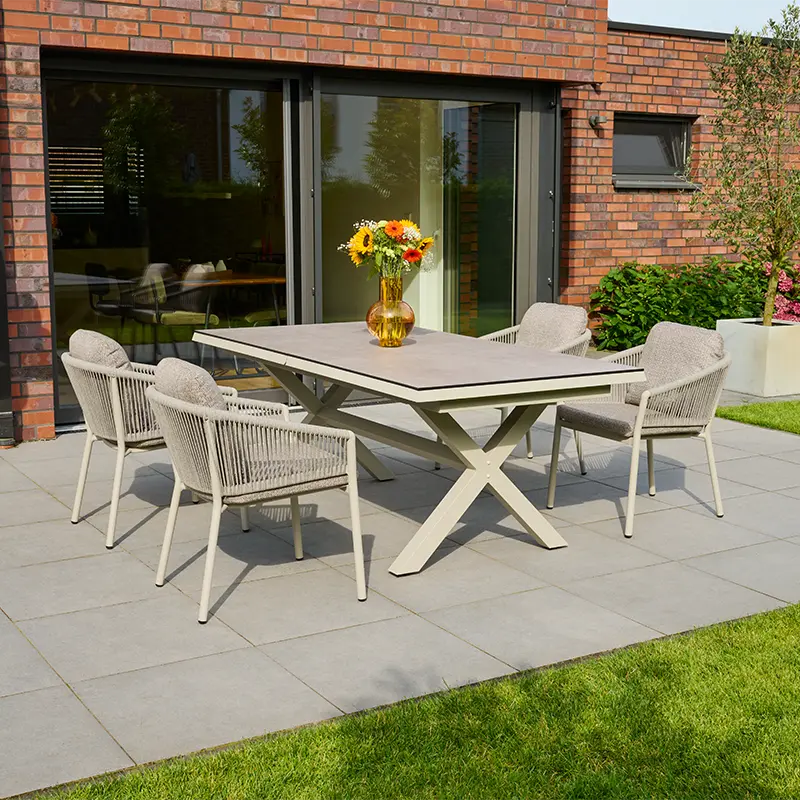 Moderner Terrassen Essbereich mit einem großen rechteckigen Tisch auf X förmigem Gestell, umgeben von sechs beigen Gartenstühlen mit geflochtenen Seiten und gepolsterten Sitzflächen. Auf dem Tisch steht eine gelbe Vase mit Sonnenblumen. Im Hintergrund befindet sich eine Backsteinfassade mit großen Schiebetüren und ein gepflegter Garten