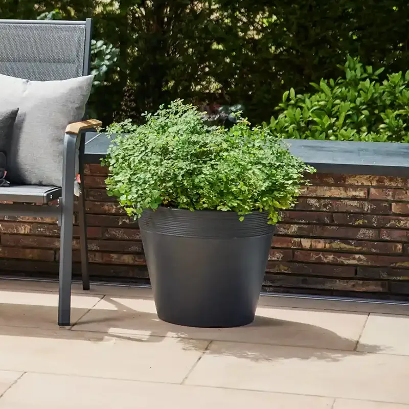 Dunkelgraues, rundes Pflanzgefäß mit grüner Bepflanzung auf einer Terrasse neben Gartenstuhl und kleiner Mauer.