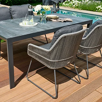 Moderne Rattanstühle mit dunklen Polstern an einem grauen Outdoor Tisch auf einer Holzterrasse am Pool.