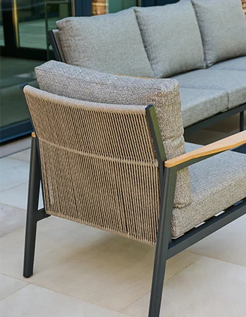 Detailaufnahme eines Outdoor‑Sessels mit geflochtener Rückenlehne, grauen Polstern und Holzarmlehnen neben einem passenden Sofa.