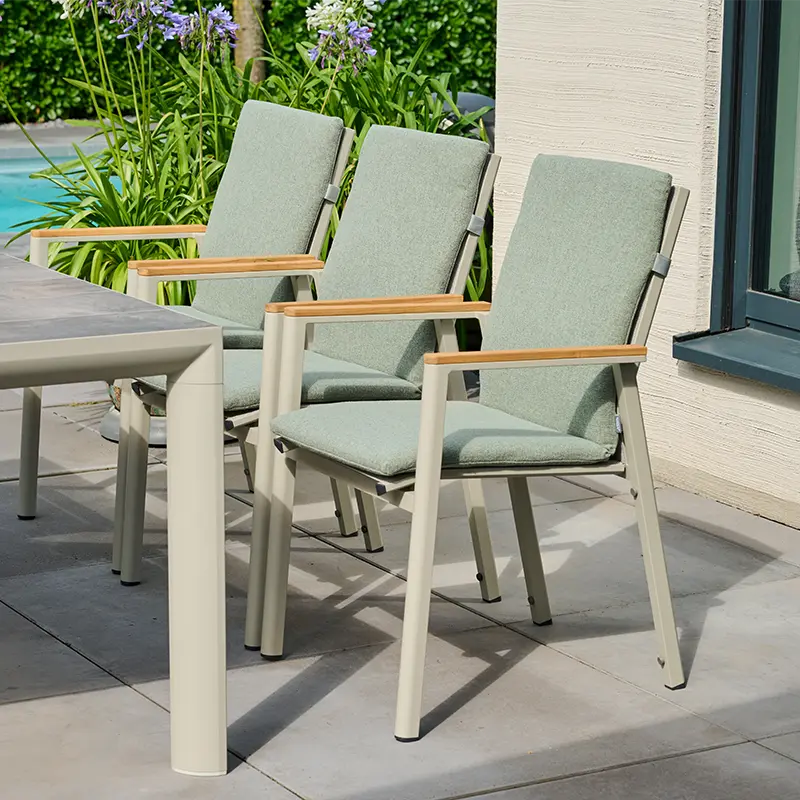 Drei Gartenstühle mit Holzarmlehnen und grünen Sitzauflagen an einem hellen Tisch auf einer Terrasse nahe einem Pool.
