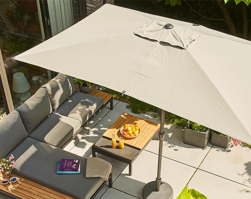 L-förmige Gartenlounge, mit Holz Ablagetisch und mit hellen Polstern unter großem, hellem rechteckigem Sonnenschirm auf sonniger Terrasse.