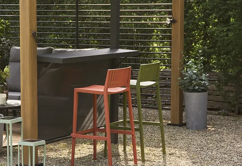 Outdoor Bar mit dunkler Theke und zwei farbigen Barhockern unter einer Pergola mit Lamellenwand.
