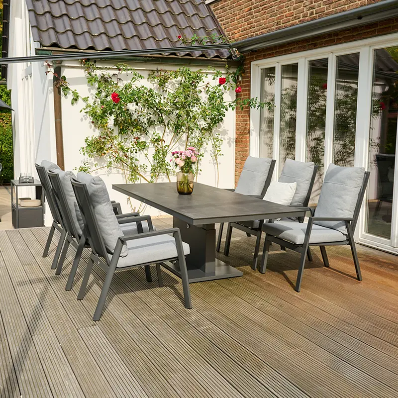 Moderner Terrassen Essbereich mit einem großen rechteckigen Tisch und mehreren gepolsterten Gartenstühlen mit grauen Bezügen. Auf dem Tisch steht eine Vase mit Blumen. Im Hintergrund wachsen Kletterrosen an der Hauswand neben großen Fenstern, und die Terrasse besteht aus Holzplanken.