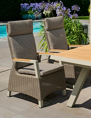 Zwei gepolsterte Garten-Hochlehner aus grauem Geflecht mit Holzarmlehnen neben einem hellen Outdoor Tisch auf einer Terrasse am Pool.