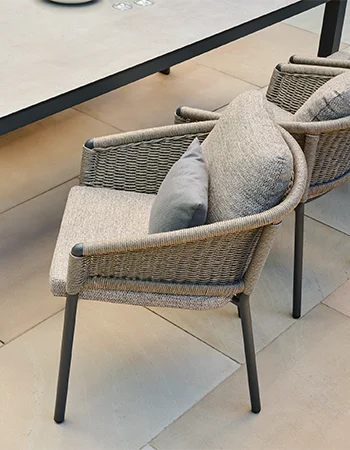 Drei moderne Gartenstühle mit hellem Metallgestell, seitlichem RopeGeflecht und beigefarbenen Polstern stehen an einem hellen  Geflecht und beigefarbenen Polstern stehen an einem hellen OutdoorTisch Tisch auf einer Terrasse, umgeben von grünen Pflanzen