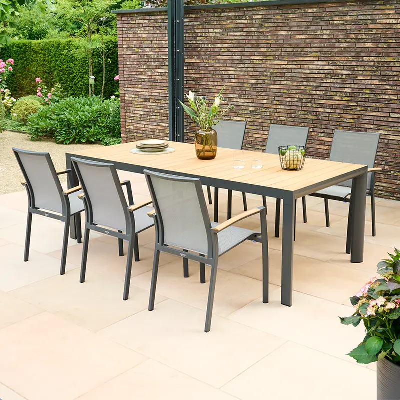 Großer rechteckiger Gartentisch mit Holzplatte und dunkelgrauem Metallgestell, umgeben von sechs Gartenstühlen mit grauer Textilbespannung und Armlehnen aus Holz. Auf dem Tisch stehen eine Vase mit Grünpflanzen, Tellerstapel, Gläser und ein Obstkorb. Die Terrasse ist von Hecken, Blumen und einer gemauerten Sichtschutzwand umgeben.