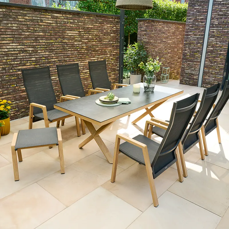 Moderne Gartensitzgruppe mit großem Tisch und sechs Hochlehnern aus Holz und Textilgewebe auf einer hellen Terrasse vor einer Backsteinwand, dekoriert mit Pflanzen.
