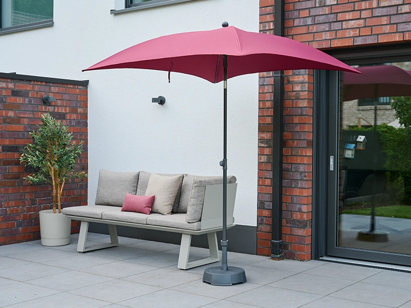 Terrassenbank mit beigefarbenen Kissen unter weinrotem Sonnenschirm vor moderner Hausfassade.