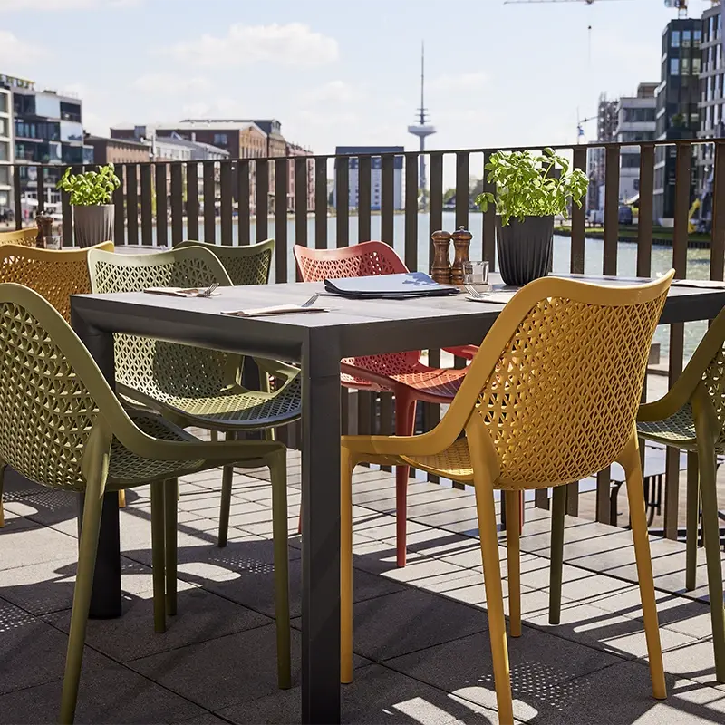 Outdoor Esstisch mit bunten Kunststoffstühlen und Pflanzen auf einer Terrasse mit Blick auf Wasser und Stadtbebauung.