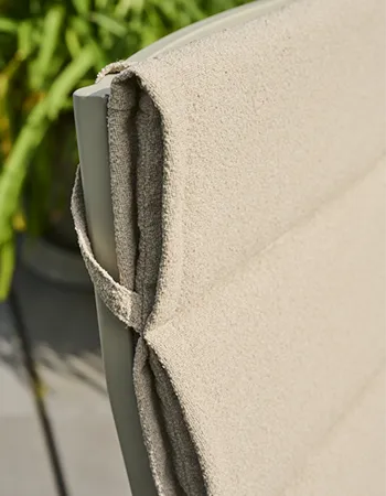 Detailaufnahme einer beigen, strukturierten Sitzauflage mit diagonalen Haltebändern auf der Rückseite an einem hellen Stuhlrahmen im Außenbereich.