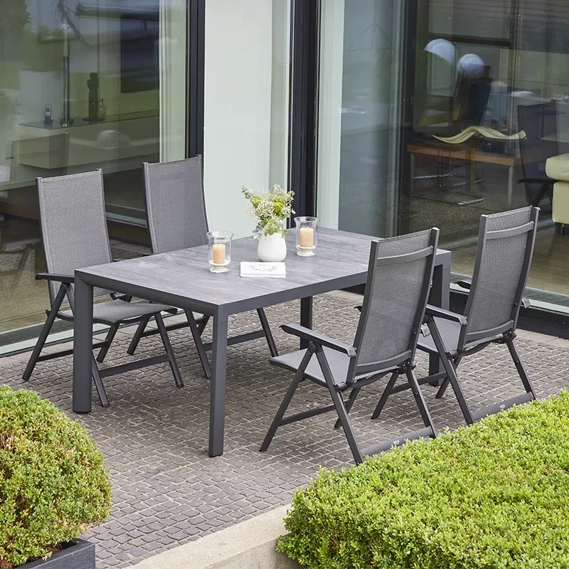 Moderner Outdoor Essbereich mit einem rechteckigen Tisch in grauer Steinoptik und fünf klappbaren Gartenstühlen mit grauer Textilbespannung. Auf dem Tisch stehen zwei Windlichter mit Kerzen und eine Vase mit Blumen. Der Bereich befindet sich vor großen Glasfronten, umgeben von gepflegten Buchshecken.