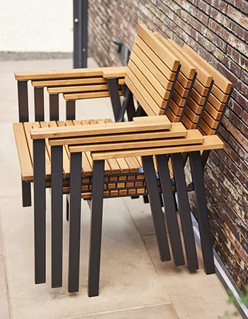 Gestapelte Gartenstühle aus Holz und schwarzem Metall auf einer Terrasse vor einer Backsteinwand. 