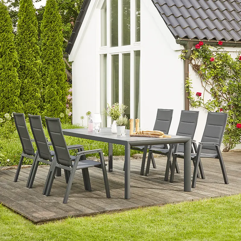 Moderner Outdoor Essbereich mit einem großen rechteckigen Tisch und mehreren Gartenstühlen mit hellblauen Polsterauflagen. Der Bereich ist von dichtem Bambus und blühenden Rhododendren umgeben. Auf dem Tisch stehen Getränke, Gebäck und eine Vase mit Blumen.