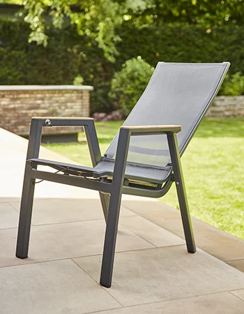 Moderner Outdoor Stuhl mit dunkelgrauem Rahmen, Holzarmlehnen und luftdurchlässiger Textilbespannung steht auf einer gefliesten Terrasse, mit grünem Garten im Hintergrund.
