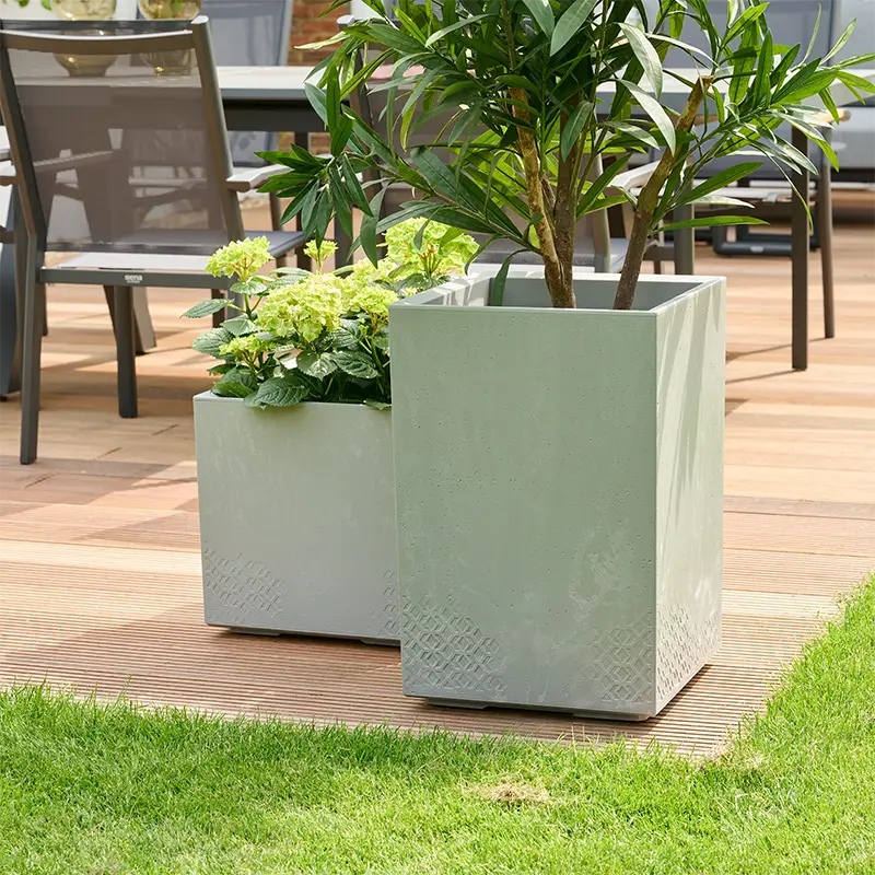 Zwei quadratische, graue Kunststoff-Pflanzgefäße in Beton-Optik mit Blühpflanzen auf einer Holzterrasse.