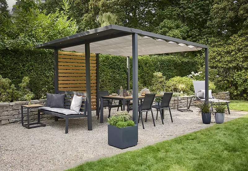 Moderne Pergola mit Stoffdach über einer Outdoor Essgruppe und Sitzbank, ergänzt durch Holz Lamellenwand und Pflanzgefäße im Garten.