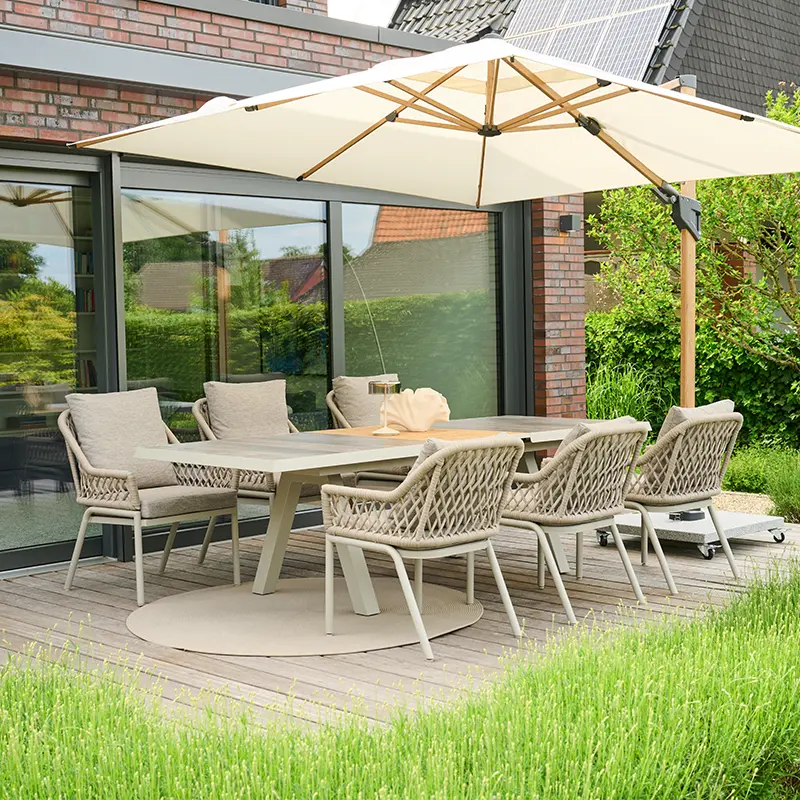Große Outdoor Essgruppe mit geflochtenen Gartenstühlen, hellem Esstisch und großem Sonnenschirm auf einer Holzterrasse vor einem modernen Haus.