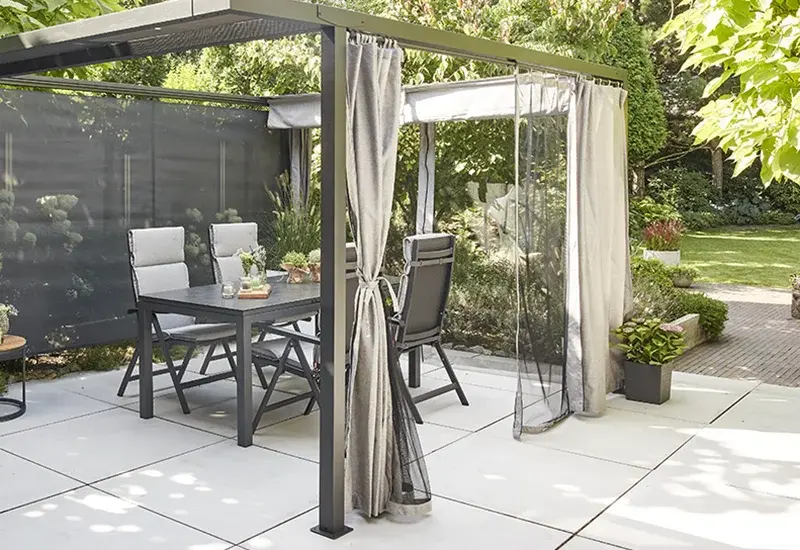 Pergola mit seitlichen Vorhängen und Textilblende über einer Outdoor Essgruppe auf einer hellen Terrassenfläche im Garten.