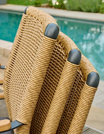 Nahaufnahme der Rückenlehnen dreier Outdoor Stühle aus Geflecht mit dunkel abgerundeten Eckkappen, vor einem Poolbereich.