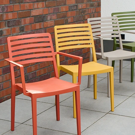 Vier farbige Kunststoffstühle in Orange, Gelb, Beige und Grün nebeneinander auf einer Terrasse vor Ziegelwand.