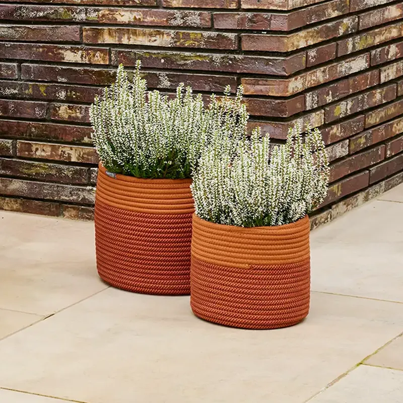 Zwei runde, bepflanzte Pflanzgefäße aus Rope in rot und orange auf heller Terrasse vor einer gemauerten Wand.