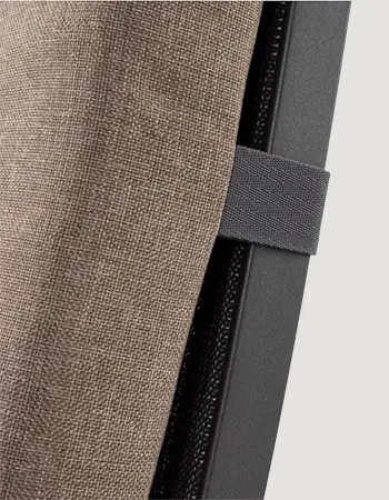 Detailaufnahme einer hellbraunen, strukturierten Sitzauflage mit elastischem Halteband an einem dunklen Metallrahmen vor hellem Hintergrund.