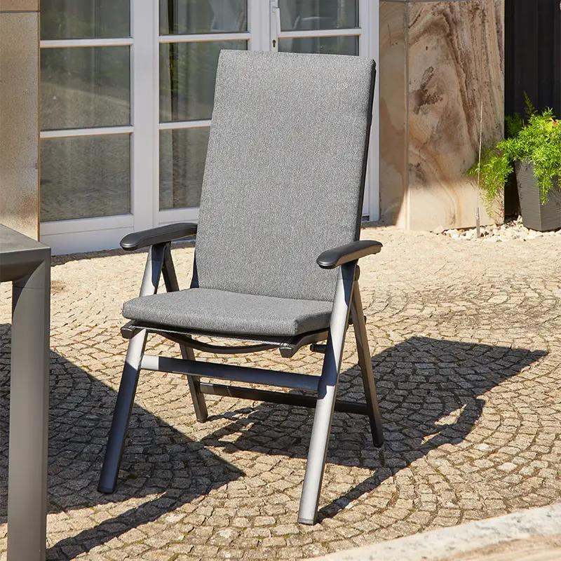 Gartenstuhl mit grauer Sitzauflage, hoher Lehne und Armlehnen auf gepflasterter Terrasse vor einer Hausfassade.