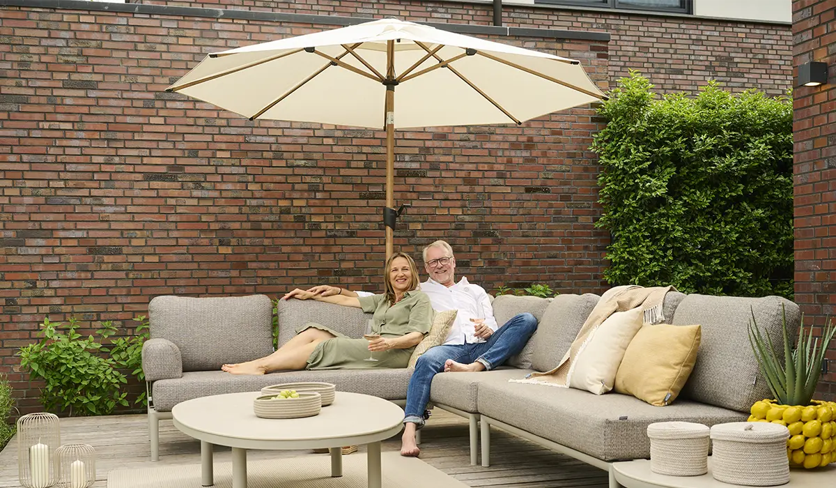 Zwei Personen entspannen auf einer modernen Outdoor Lounge unter einem großen Mittelstockschirm auf einer Terrasse mit Backsteinwand und Pflanzen.