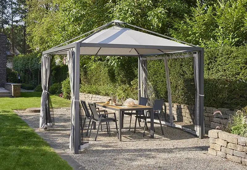 Pavillon mit Spitzdach und grauem Stoffdach, zurückgezogenen Vorhängen an den Seiten über einer Outdoor Essgruppe im Garten, umgeben von Steinmauern und Grünpflanzen.