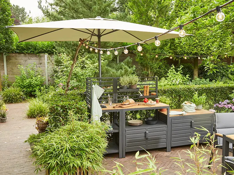 Outdoor-Küchenmodul aus dunkelgrauem Holz mit Arbeitsfläche unter großem beigem Sonnenschirm, umgeben von dichtem Grün und Dekolichterkette.
