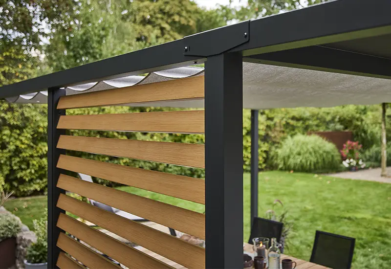 Lamellenwand in Holzoptik unter einem hellen, textilen Pergola-Stoffdach mit Blick auf dichte Gartenbepflanzung.