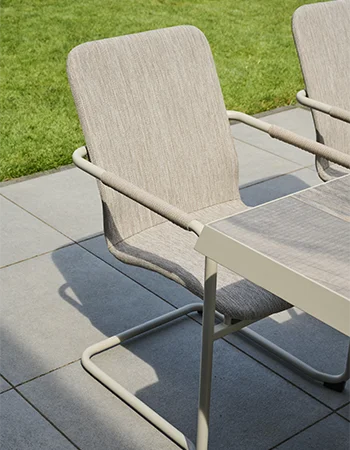 Outdoor Stuhl mit hellem, gepolstertem Bezug und leicht geschwungenem Metallgestell steht neben einem Gartentisch auf einer gefliesten Terrasse, mit Rasenfläche im Hintergrund.