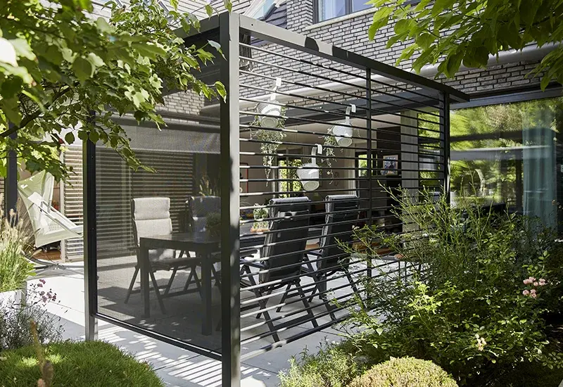 Pergola in anthrazit mit seitlichem Aluminium Lamellenpanel  auf einer Terrasse mit Gartenmöbeln und umgebender Bepflanzung.