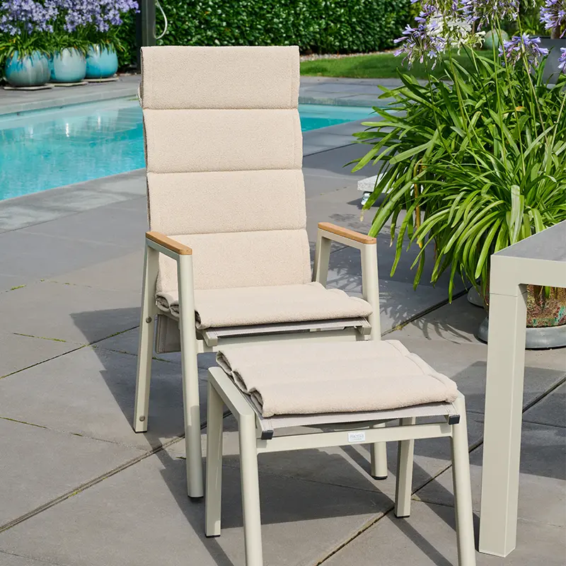 Gartensessel in matt seidengrau mit passender Auflage mit Steppnähten und Hocker auf heller Terrasse neben Pflanzen und Pool.