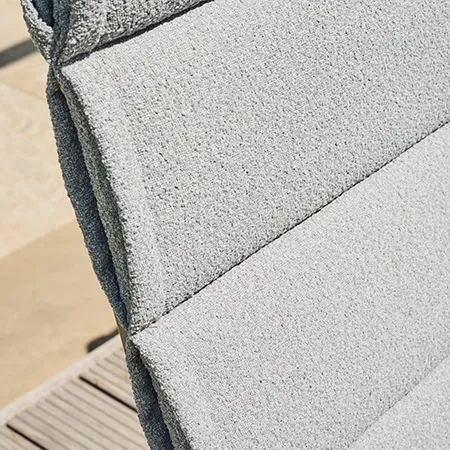 Detailaufnahme einer hellgrauen, strukturierten Polsterauflage mit horizontalen Steppnähten im Sonnenlicht vor unscharfem Außenbereich.