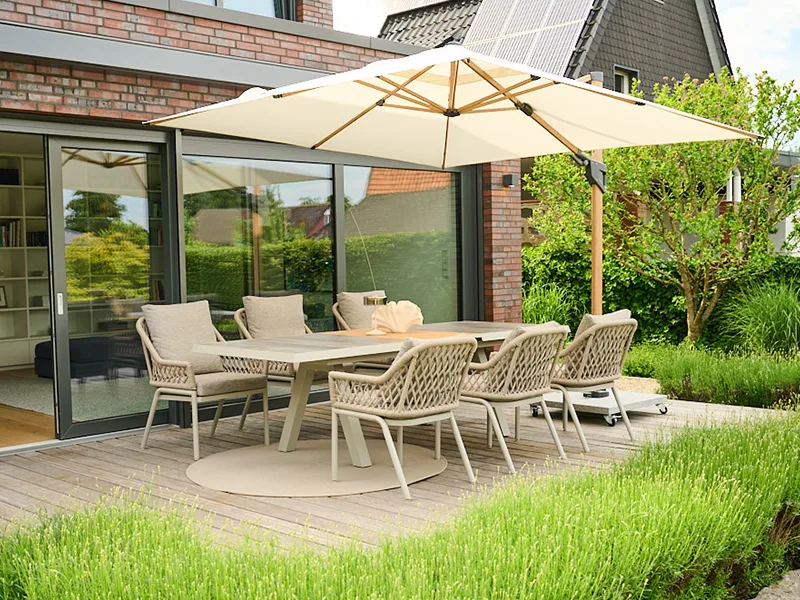 Großer Esstisch mit geflochtenen Stühlen unter hellem Ampelschirm auf Holzterrasse vor moderner Fassade und Grünpflanzen.