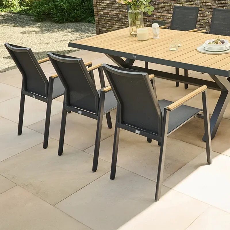 Gartenessbereich mit einem rechteckigen Tisch aus Holz und Metall, umgeben von mehreren dunkelgrauen Stühlen und Relaxsessel Hochlehnern. Auf dem Tisch stehen Wassergläser, ein Teller mit Trauben und zwei Vasen mit Blumen. Im Hintergrund befinden sich eine gemauerte Sichtschutzwand, Bäume und ein gepflegter Gartenweg