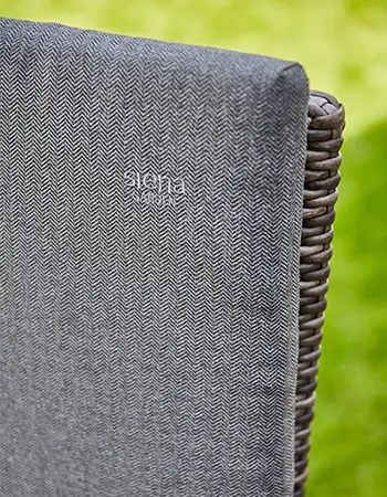 Detailaufnahme einer grau gepolsterten Sitzauflage mit feinem Fischgrätmuster und dezent gesticktem SIENA GARDEN Logo vor unscharfem grünem Hintergrund.