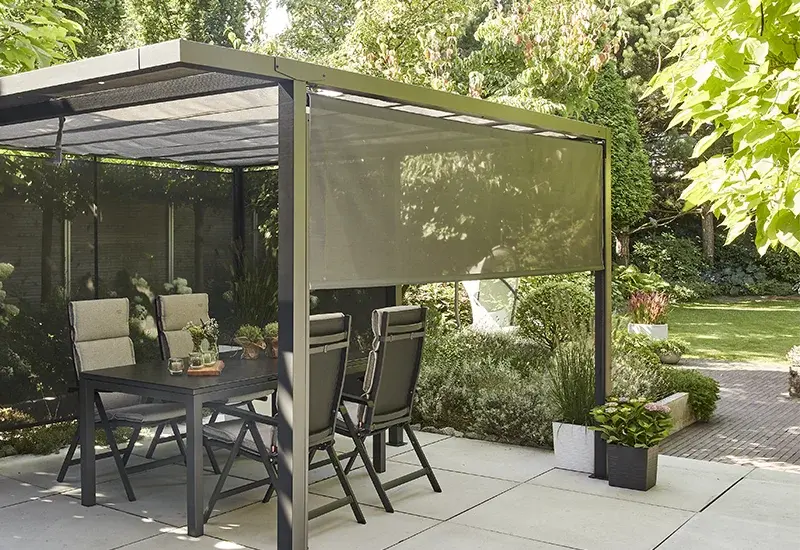 Pergola mit halb ausgefahrener, hellgrauer Textil Sonnenblende über einer Outdoor Essgruppe im Garten.