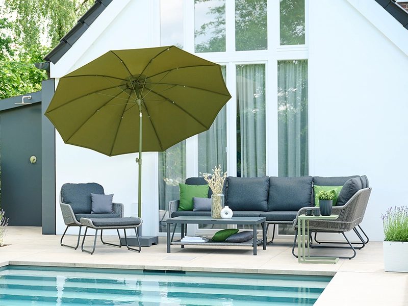 Graue Gartenlounge und Sessel mit grünen Dekokissen unter olivgrünem Sonnenschirm am Pool vor weißem Haus.