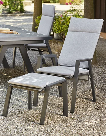 Gepolsterter Gartenstuhl mit passendem Fußhocker steht auf einem Kiesuntergrund neben einem großen Outdoor Esstisch. Die Möbel haben einen dunkelgrauen Metallrahmen und helle Polster. Im Hintergrund sind weitere Gartenstühle und grüne Pflanzen zu sehen