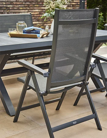 Gartenstuhl mit dunkelgrauem Metallrahmen und feinmaschiger Textilbespannung steht an einem großen Outdoor Tisch. Auf dem Tisch liegen ein Tablett mit Geschirr, ein Buch und eine Vase mit Blumen.