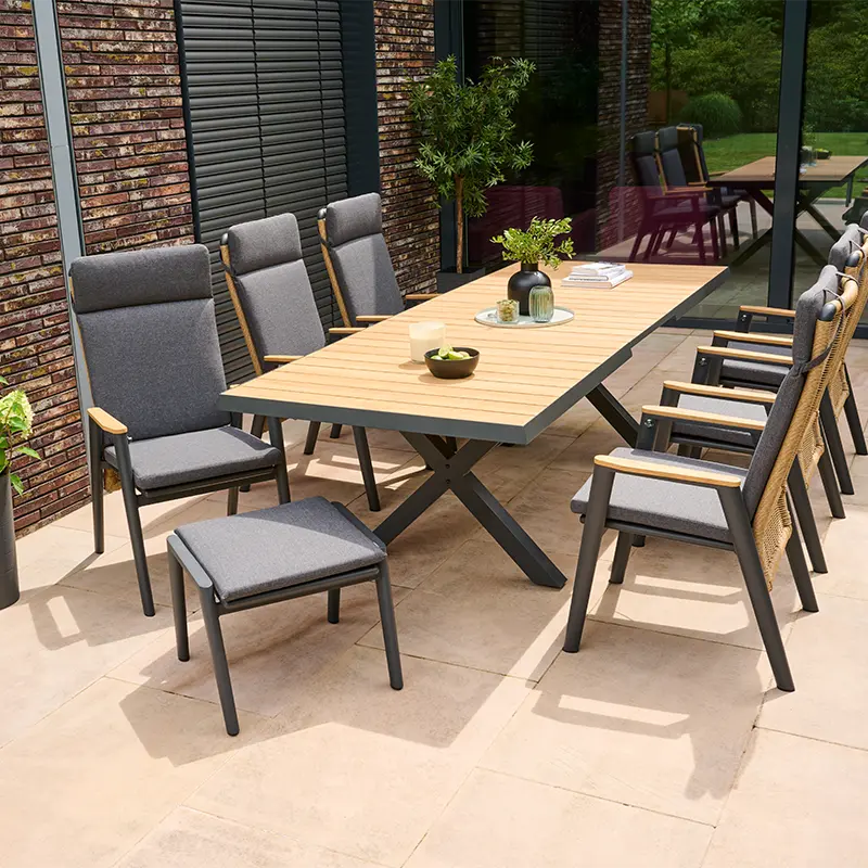 Großer Terrassentisch mit hellen Holzlamellen und dunkelgrauem Metallgestell, umgeben von mehreren gepolsterten Gartenstühlen mit Holzarmlehnen und geflochtener Rückenlehne. Auf dem Tisch stehen Deko-Objekte und Pflanzen. Die Terrasse liegt vor einer modernen Hausfassade mit bodentiefen Fenstern