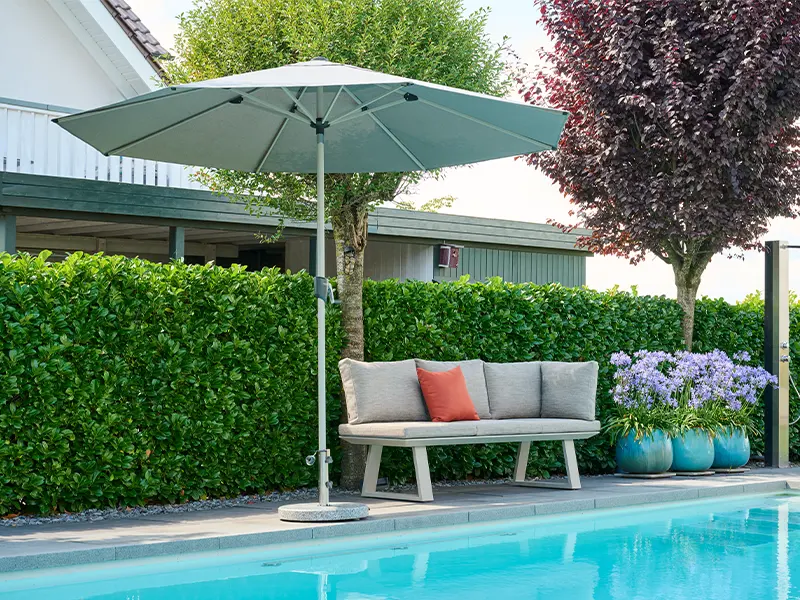 Helle Gartenbank mit Kissen unter grauem Sonnenschirm am Pool, umgeben von grüner Hecke und türkisen Pflanzkübeln.
