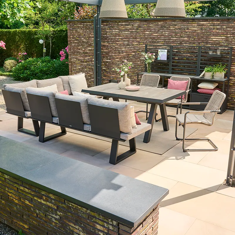 Modern gestalteter Terrassenbereich mit einem großen rechteckigen Esstisch, einer langen Gartenbank mit vielen Kissen und zwei leichten Metallstühlen. Auf dem Tisch stehen dekorative Vasen mit Blumen. Im Hintergrund befinden sich eine gemauerte Sichtschutzwand, Pflanzenregale mit Deko und ein gepflegter Garten.